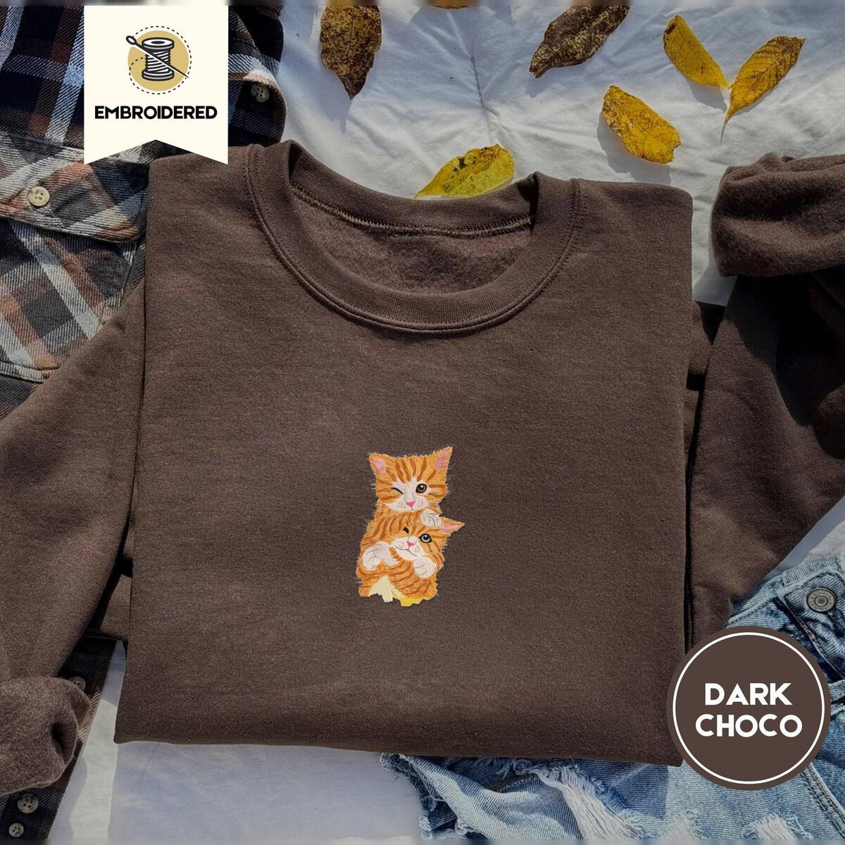 Kitten Sweatshirt – Dark Chocolate Dark Chocolate crewneck sweatshirt on a plaid flannel shirt with yellow autumn leaves and denim shorts, embroidered with two fluffy orange tabby kittens, one winking with a paw on the other's head
