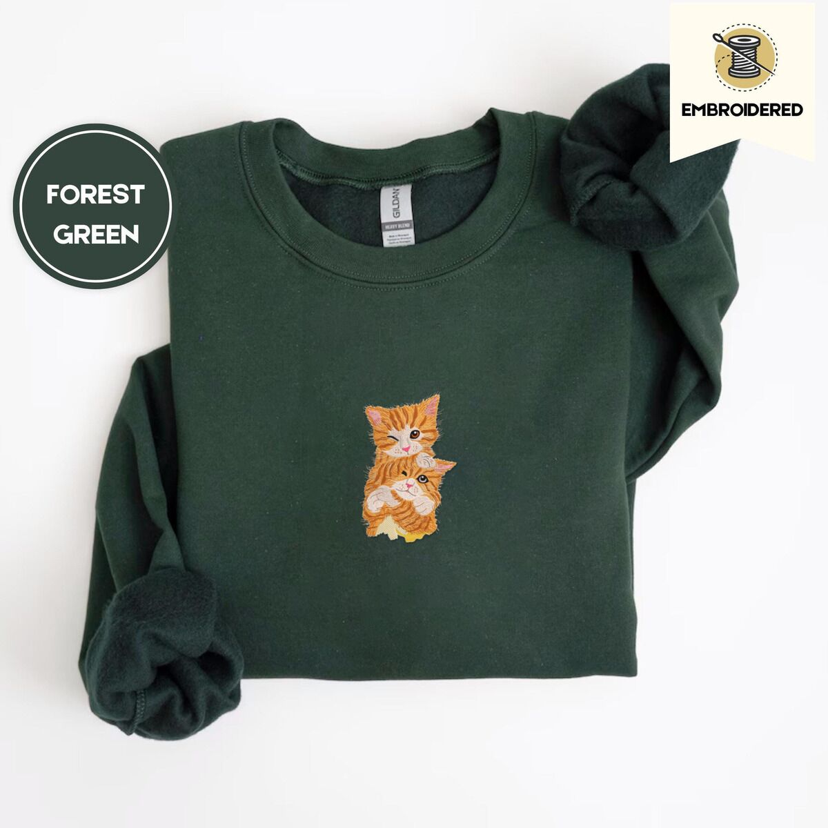 Kitten Sweatshirt – Forest Green Forest Green crewneck sweatshirt folded on a white background, embroidered with two fluffy orange tabby kittens, one winking with a paw on the other's head