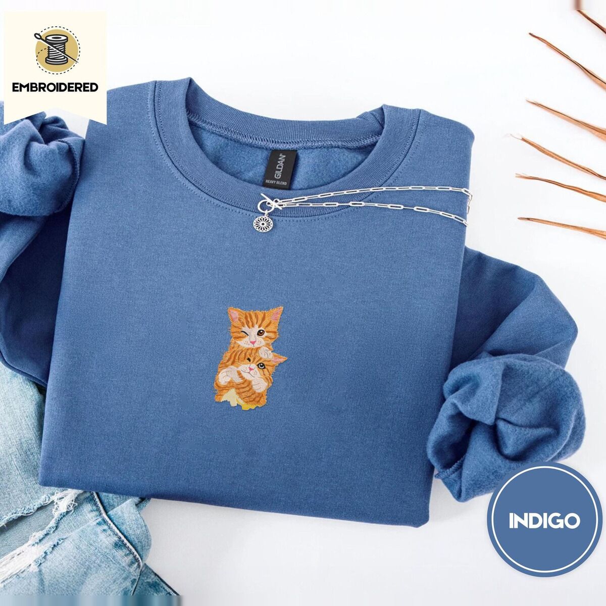 Kitten Sweatshirt – Indigo Blue Indigo Blue crewneck sweatshirt folded with light jeans and dried twigs, embroidered with two fluffy orange tabby kittens, one winking with a paw on the other's head