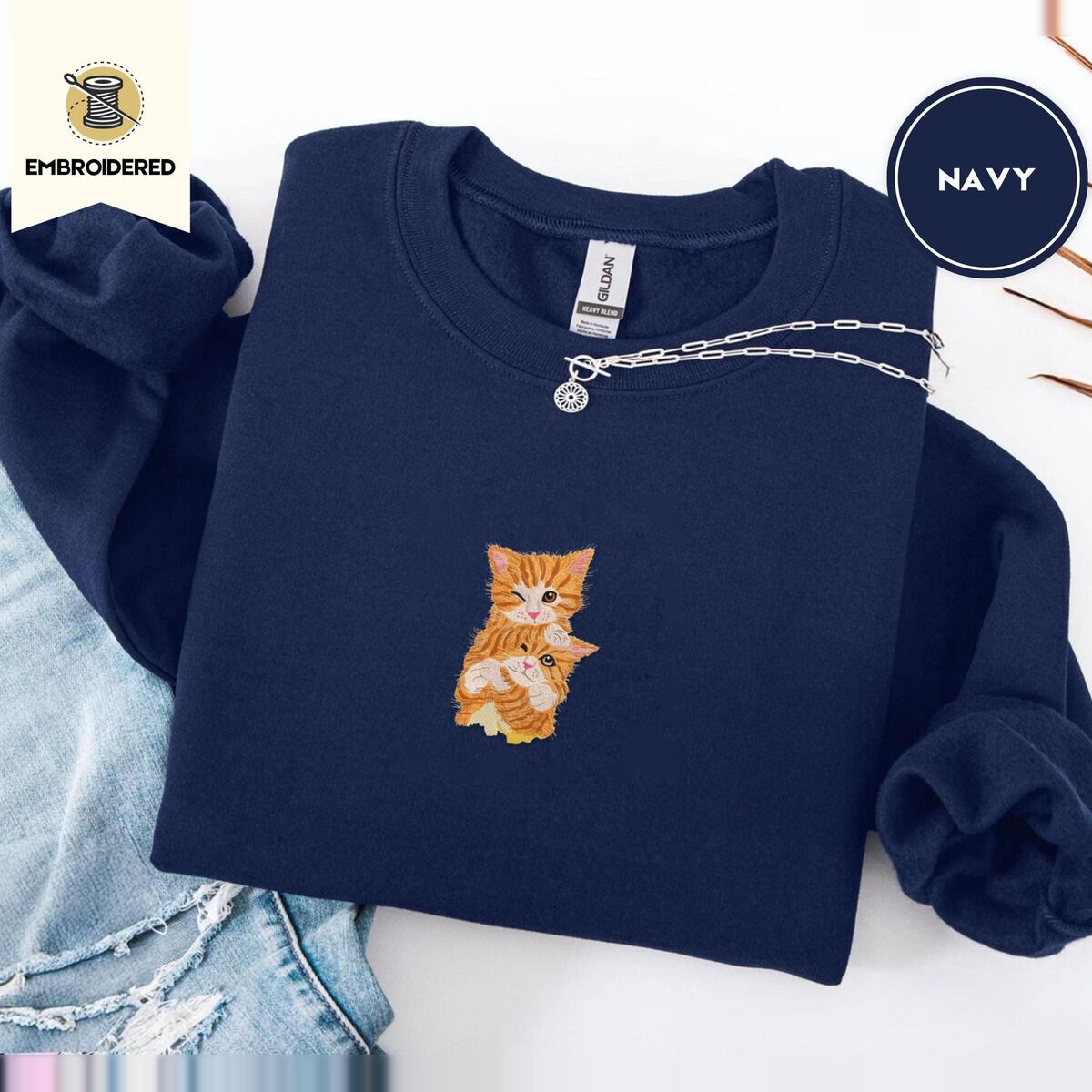 Kitten Sweatshirt – Navy Navy crewneck sweatshirt folded with ripped jeans and a silver chain necklace, embroidered with two fluffy orange tabby kittens, one winking with a paw on the other's head