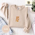 Sand crewneck sweatshirt folded with denim shorts, white sneakers and a potted plant, embroidered with two fluffy orange tabby kittens, one winking with a paw on the other's head