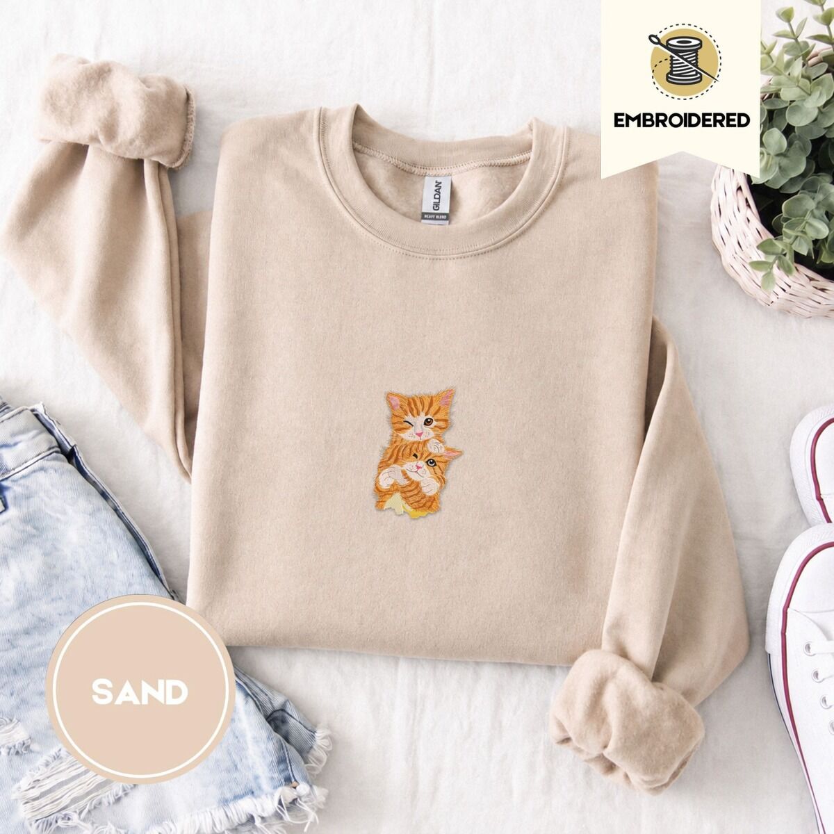 Kitten Sweatshirt – Sand Sand crewneck sweatshirt folded with denim shorts, white sneakers and a potted plant, embroidered with two fluffy orange tabby kittens, one winking with a paw on the other's head