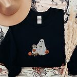 Black crewneck sweatshirt laid flat with a brown hat and pampas grass, embroidered with a ghost lab mom holding a coffee cup and a jack-o'-lantern pail beside a tan lab in a ghost sheet with its own pumpkin pail