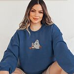 Smiling woman sitting in a blue crewneck sweatshirt embroidered with a ghost lab mom holding a coffee cup and a jack-o'-lantern pail beside a tan lab in a ghost sheet with its own pumpkin pail