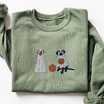 Green crewneck sweatshirt laid flat on a white background, embroidered with two Labrador ghosts: a light cream lab draped in a white sheet and a black lab in a sheet holding a jack-o'-lantern, with a pumpkin between them