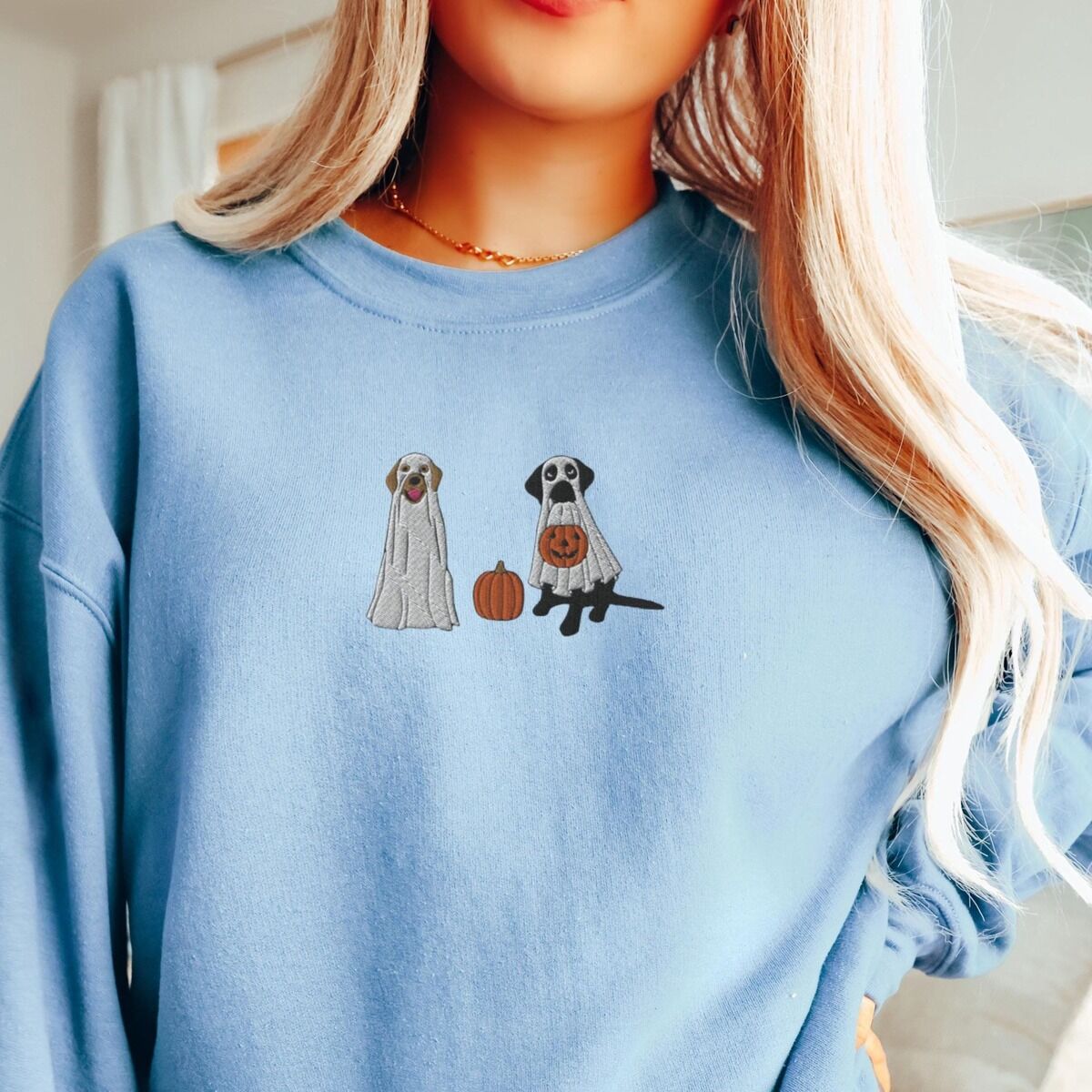 Labrador Ghosts Sweatshirt - worn Woman with long blonde hair in a light blue crewneck sweatshirt embroidered with two Labrador ghosts: a light cream lab draped in a white sheet and a black lab in a sheet holding a jack-o'-lantern, with a pumpkin between them