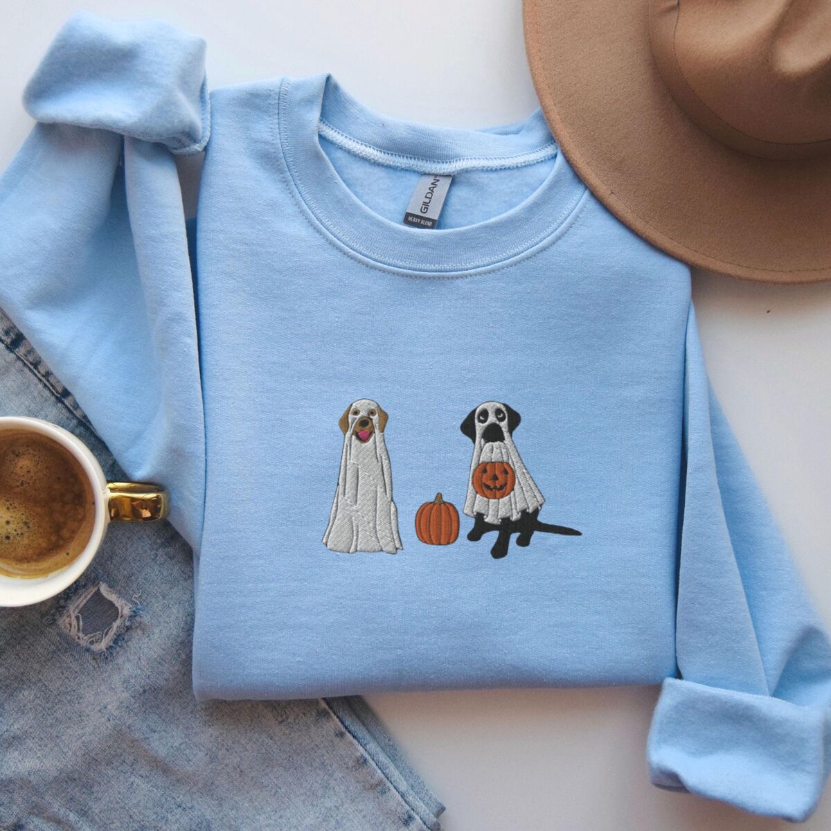 Labrador Ghosts Sweatshirt - Light Blue Light Blue crewneck sweatshirt laid flat beside jeans, a coffee cup and a brown hat, embroidered with two Labrador ghosts: a light cream lab draped in a white sheet and a black lab in a sheet holding a jack-o'-lantern, with a pumpkin between them