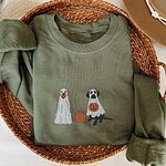 Military Green crewneck sweatshirt laid flat in a woven basket with a brown hat, embroidered with two Labrador ghosts: a light cream lab draped in a white sheet and a black lab in a sheet holding a jack-o'-lantern, with a pumpkin between them