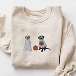 Sand crewneck sweatshirt laid flat on a white background, embroidered with two Labrador ghosts: a light cream lab draped in a white sheet and a black lab in a sheet holding a jack-o'-lantern, with a pumpkin between them