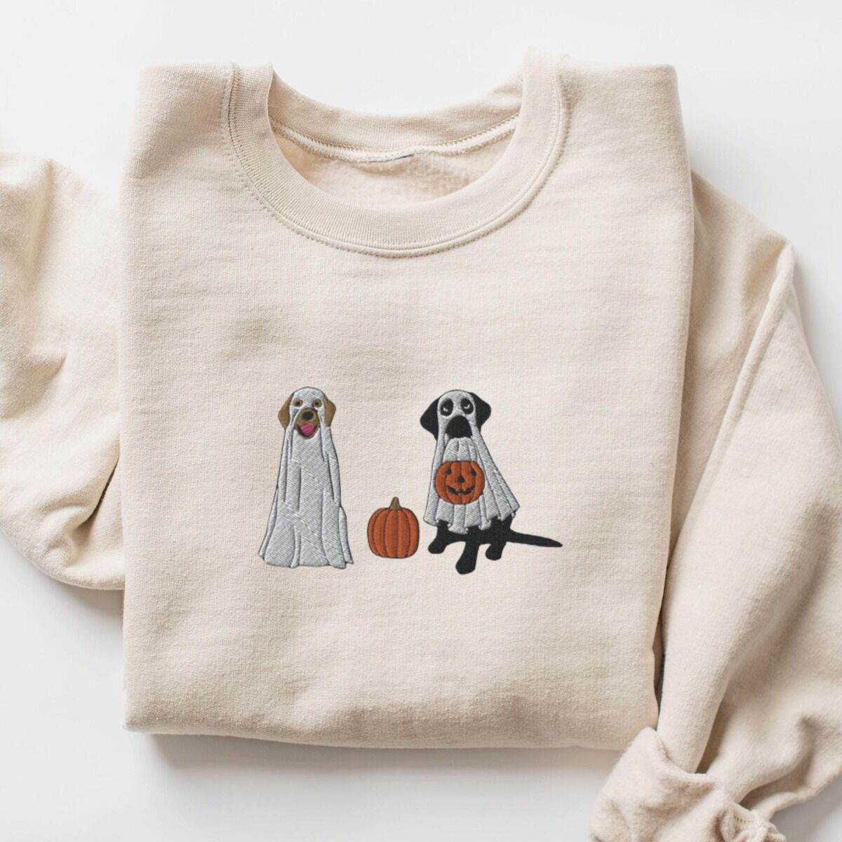 Labrador Ghosts Sweatshirt – Sand Sand crewneck sweatshirt laid flat on a white background, embroidered with two Labrador ghosts: a light cream lab draped in a white sheet and a black lab in a sheet holding a jack-o'-lantern, with a pumpkin between them