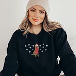 Woman in a knit beanie and a Black crewneck sweatshirt embroidered with a sitting Labrador in a red Santa hat and a fringed red scarf, framed by snowflakes