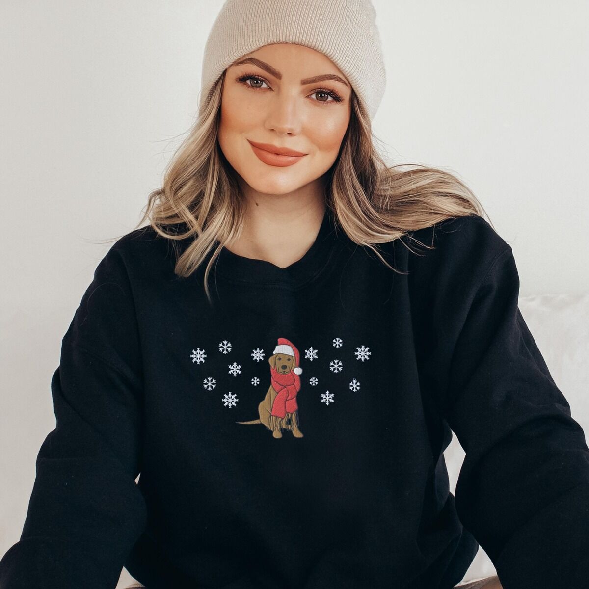 Labrador Snowflake Christmas Sweatshirt – Black Woman in a knit beanie and a Black crewneck sweatshirt embroidered with a sitting Labrador in a red Santa hat and a fringed red scarf, framed by snowflakes