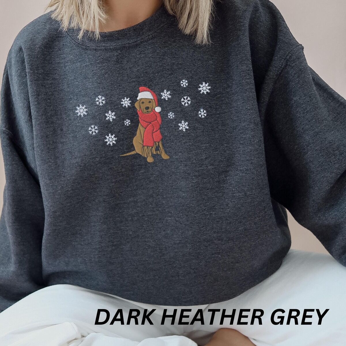 Labrador Snowflake Christmas Sweatshirt – Dark Heather Woman sitting in a Dark Heather crewneck sweatshirt embroidered with a sitting Labrador in a red Santa hat and a fringed red scarf, framed by snowflakes