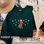 Forest Green crewneck sweatshirt laid flat with a hat and dried pampas, embroidered with a sitting Labrador in a red Santa hat and a fringed red scarf, framed by snowflakes