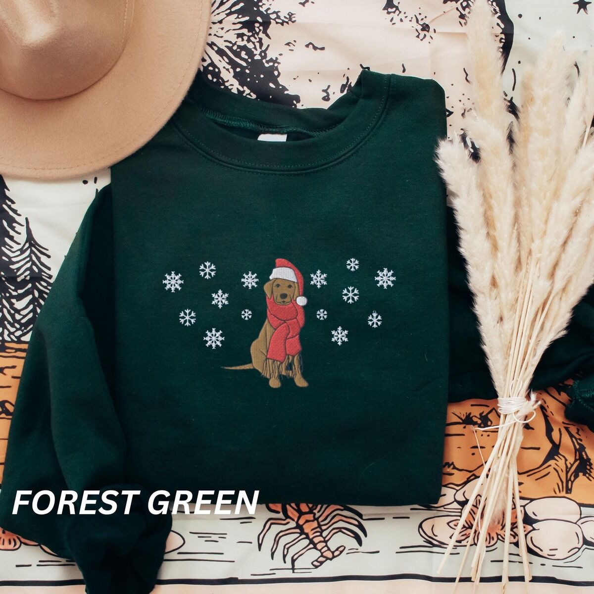 Labrador Snowflake Christmas Sweatshirt – Forest Green Forest Green crewneck sweatshirt laid flat with a hat and dried pampas, embroidered with a sitting Labrador in a red Santa hat and a fringed red scarf, framed by snowflakes