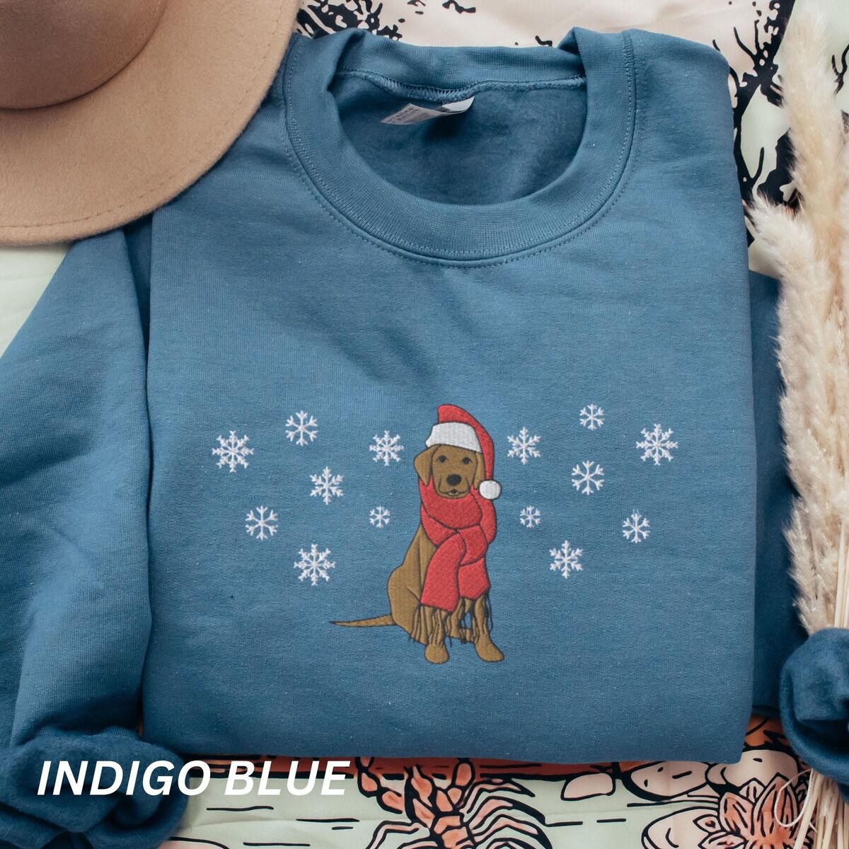 Labrador Snowflake Christmas Sweatshirt – Indigo Blue Indigo Blue crewneck sweatshirt laid flat with a hat and dried pampas, embroidered with a sitting Labrador in a red Santa hat and a fringed red scarf, framed by snowflakes