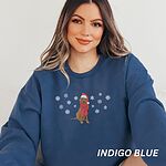 Smiling woman sitting in an Indigo Blue crewneck sweatshirt embroidered with a sitting Labrador in a red Santa hat and a fringed red scarf, framed by snowflakes