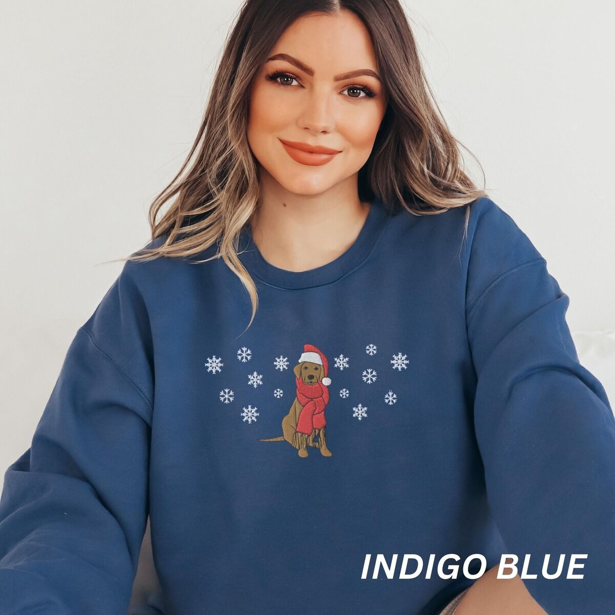 Labrador Snowflake Christmas Sweatshirt - worn Smiling woman sitting in an Indigo Blue crewneck sweatshirt embroidered with a sitting Labrador in a red Santa hat and a fringed red scarf, framed by snowflakes