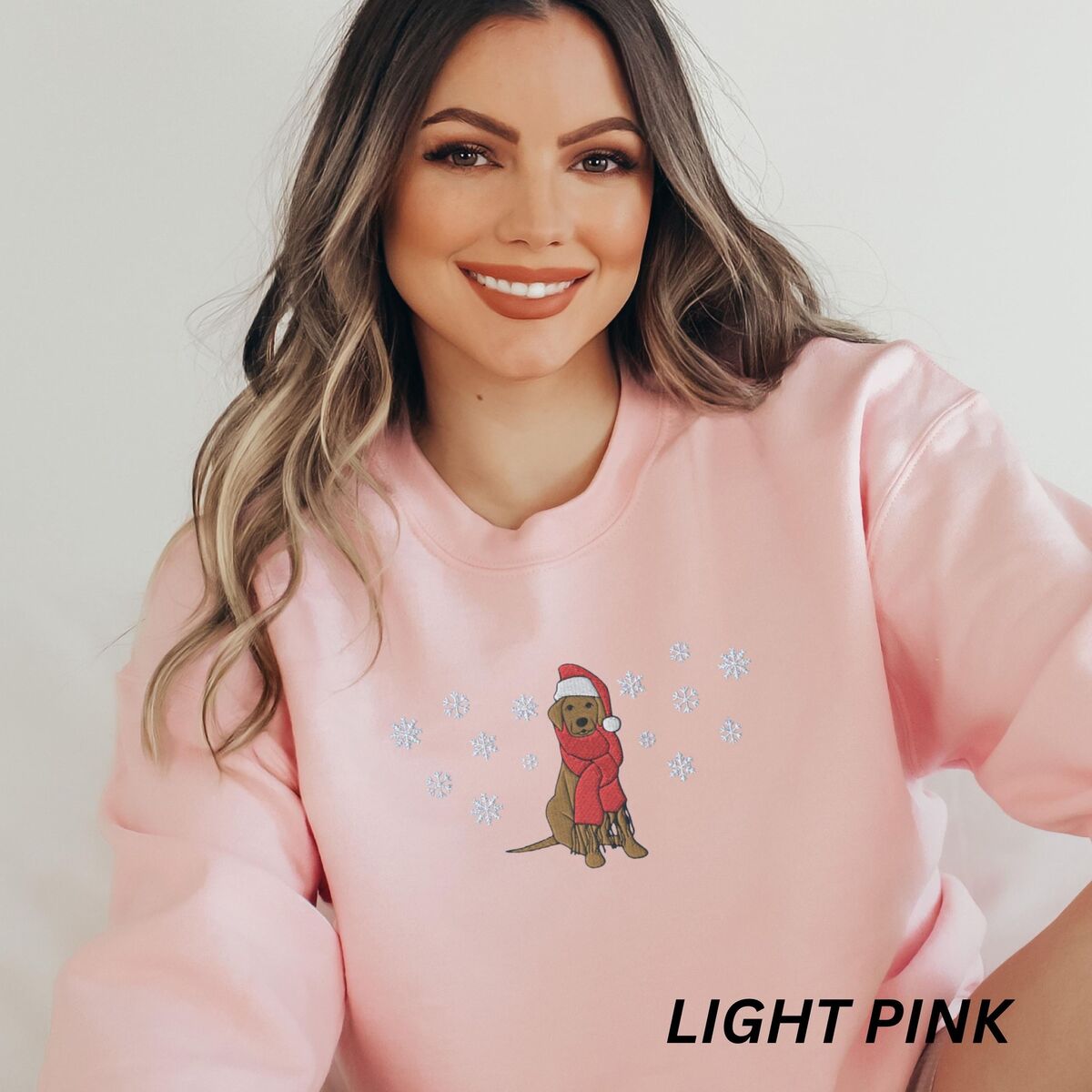 Labrador Snowflake Christmas Sweatshirt - Light Pink Smiling woman in a Light Pink crewneck sweatshirt embroidered with a sitting Labrador in a red Santa hat and a fringed red scarf, framed by snowflakes