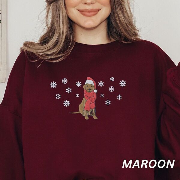 Smiling woman in a Maroon crewneck sweatshirt embroidered with a sitting Labrador in a red Santa hat and a fringed red scarf, framed by snowflakes