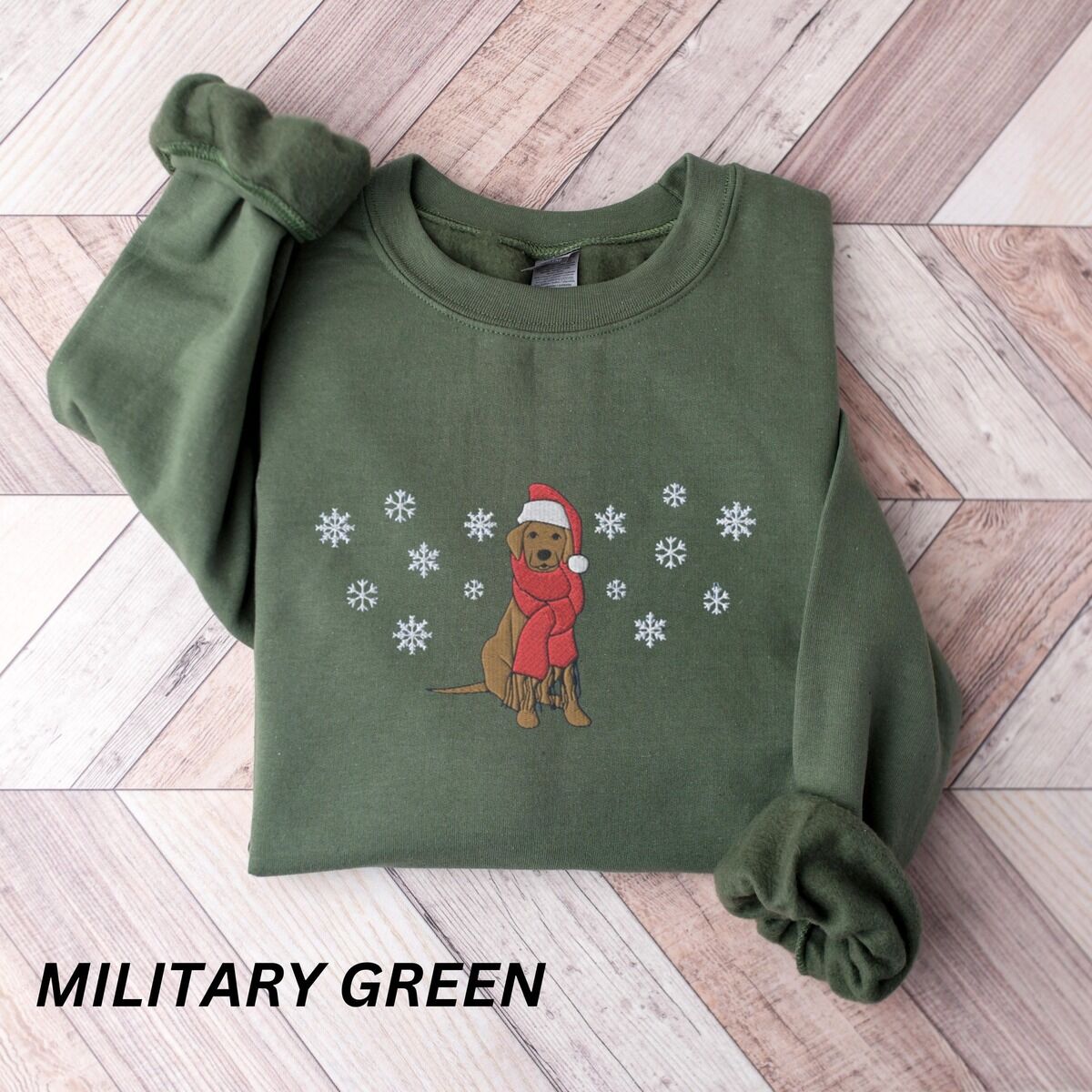 Labrador Snowflake Christmas Sweatshirt – Military Green Military Green crewneck sweatshirt laid flat on a chevron wood floor, embroidered with a sitting Labrador in a red Santa hat and a fringed red scarf, framed by snowflakes