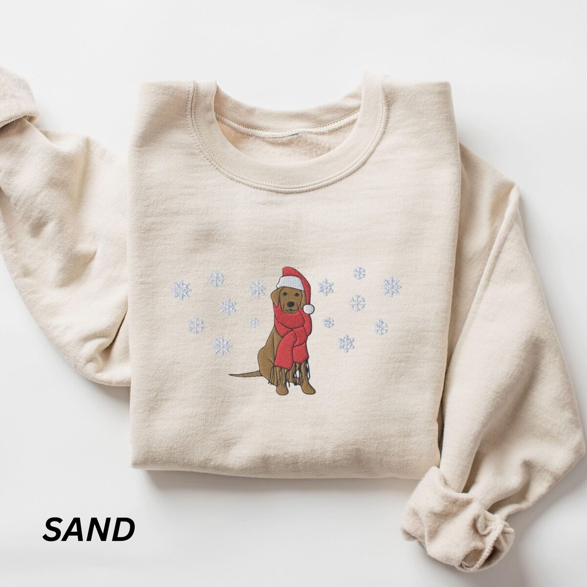 Labrador Snowflake Christmas Sweatshirt – Sand Sand crewneck sweatshirt laid flat with rolled sleeves, embroidered with a sitting Labrador in a red Santa hat and a fringed red scarf, framed by snowflakes