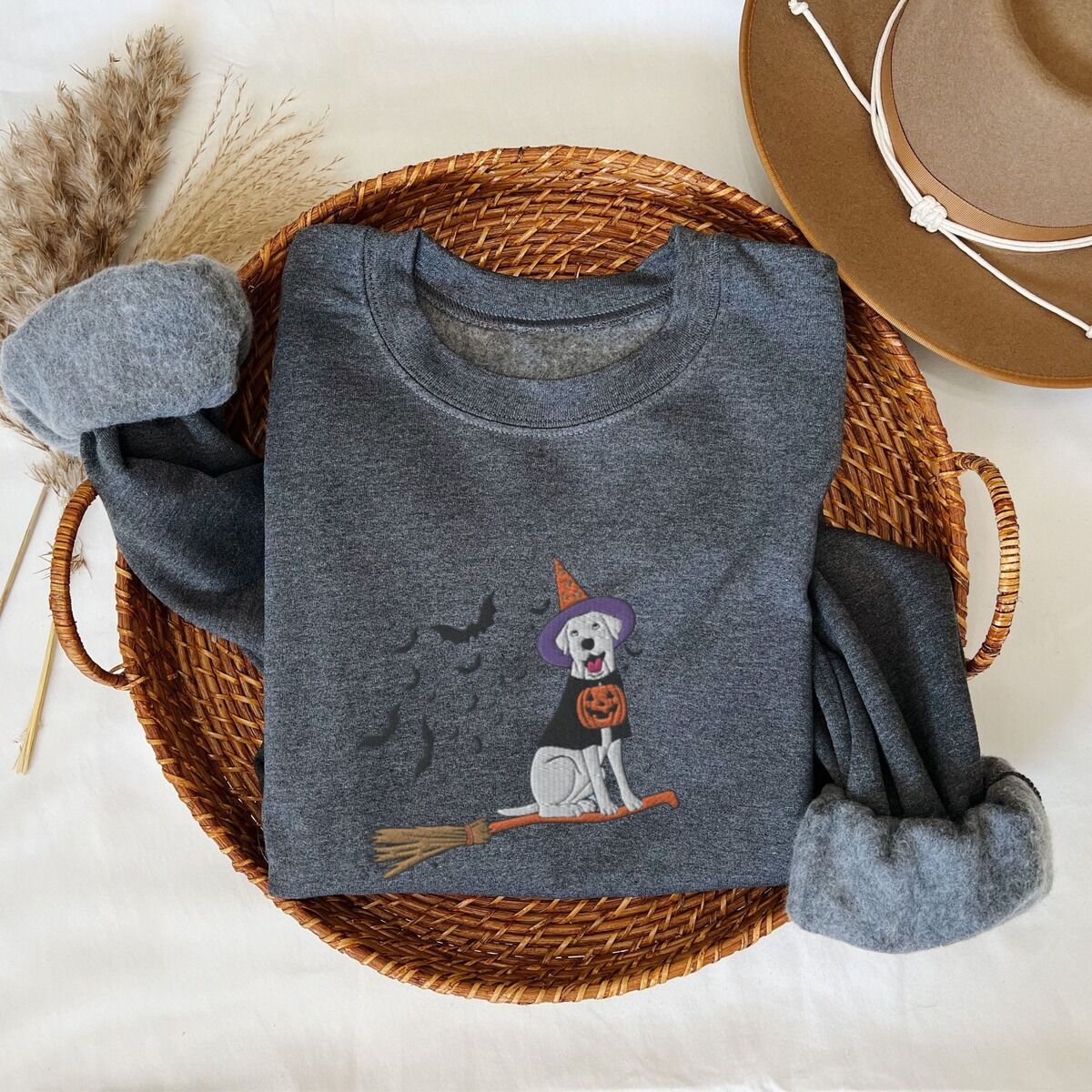 Labrador Witch Sweatshirt – Dark Heather Dark Heather crewneck sweatshirt laid flat in a woven basket with a brown hat, embroidered with a pale cream lab in a purple and orange witch hat and a jack-o'-lantern cape, riding a broomstick with a trail of bats