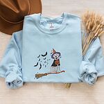 Light Blue crewneck sweatshirt laid flat with a brown hat and wheat stems, embroidered with a pale cream lab in a purple and orange witch hat and a jack-o'-lantern cape, riding a broomstick with a trail of bats