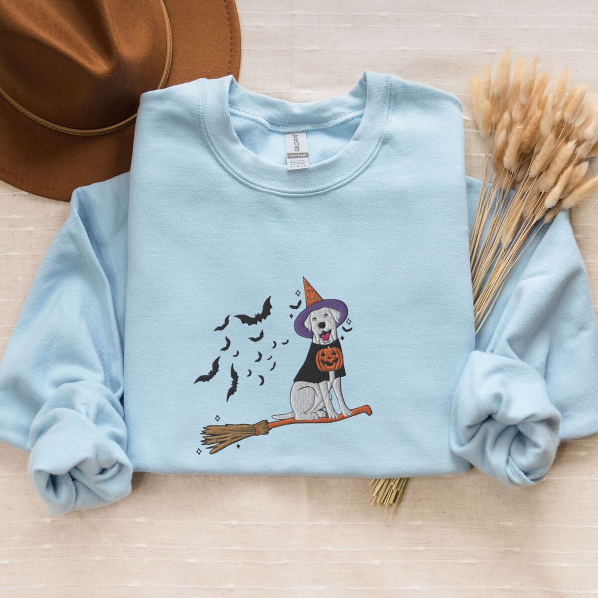 Labrador Witch Sweatshirt - Light Blue Light Blue crewneck sweatshirt laid flat with a brown hat and wheat stems, embroidered with a pale cream lab in a purple and orange witch hat and a jack-o'-lantern cape, riding a broomstick with a trail of bats