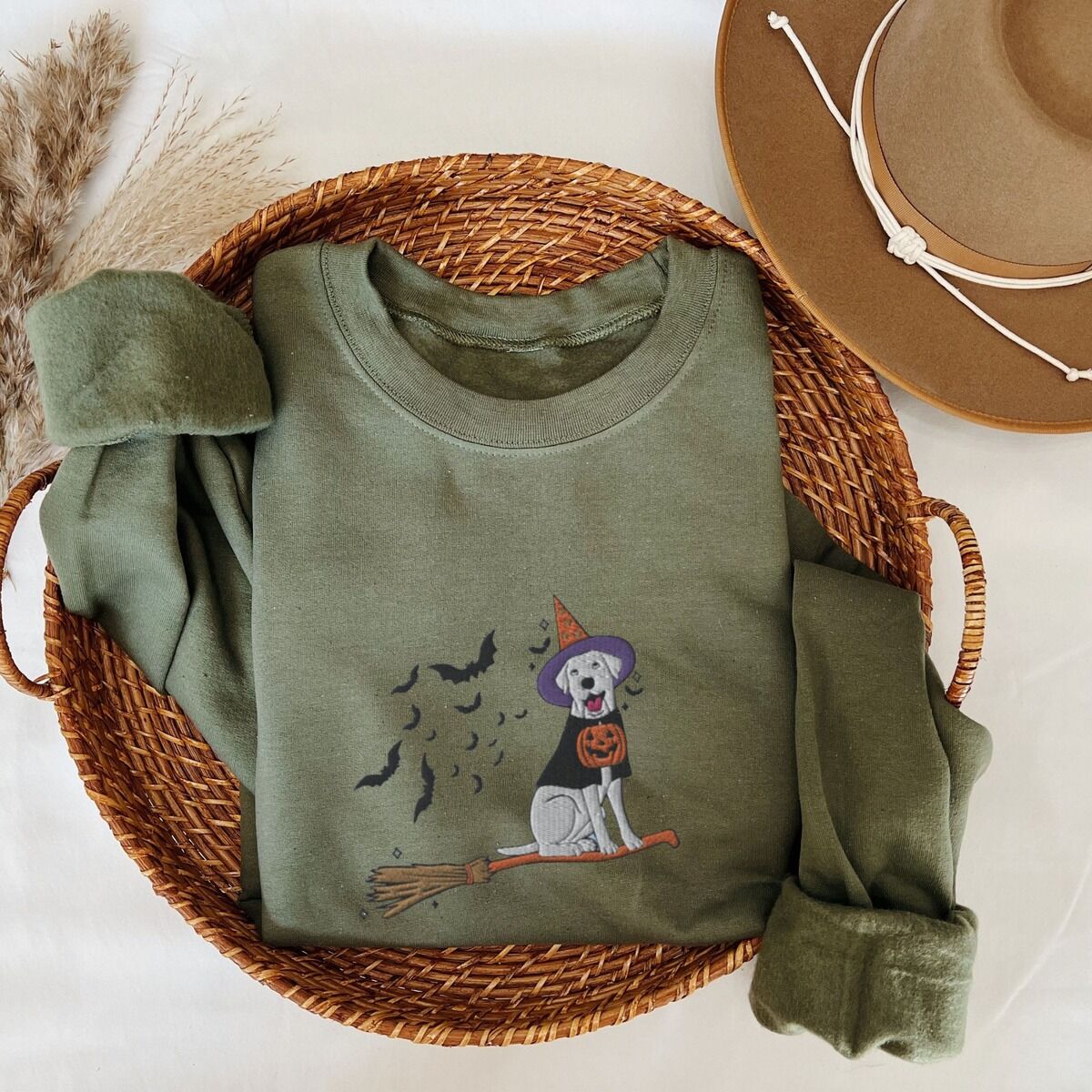 Labrador Witch Sweatshirt – Military Green Military Green crewneck sweatshirt laid flat in a woven basket with a brown hat, embroidered with a pale cream lab in a purple and orange witch hat and a jack-o'-lantern cape, riding a broomstick with a trail of bats