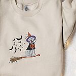 Sand crewneck sweatshirt laid flat with a sleeve folded over, embroidered with a pale cream lab in a purple and orange witch hat and a jack-o'-lantern cape, riding a broomstick with a trail of bats