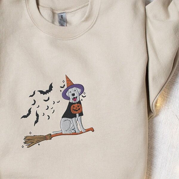 Sand crewneck sweatshirt laid flat with a sleeve folded over, embroidered with a pale cream lab in a purple and orange witch hat and a jack-o'-lantern cape, riding a broomstick with a trail of bats