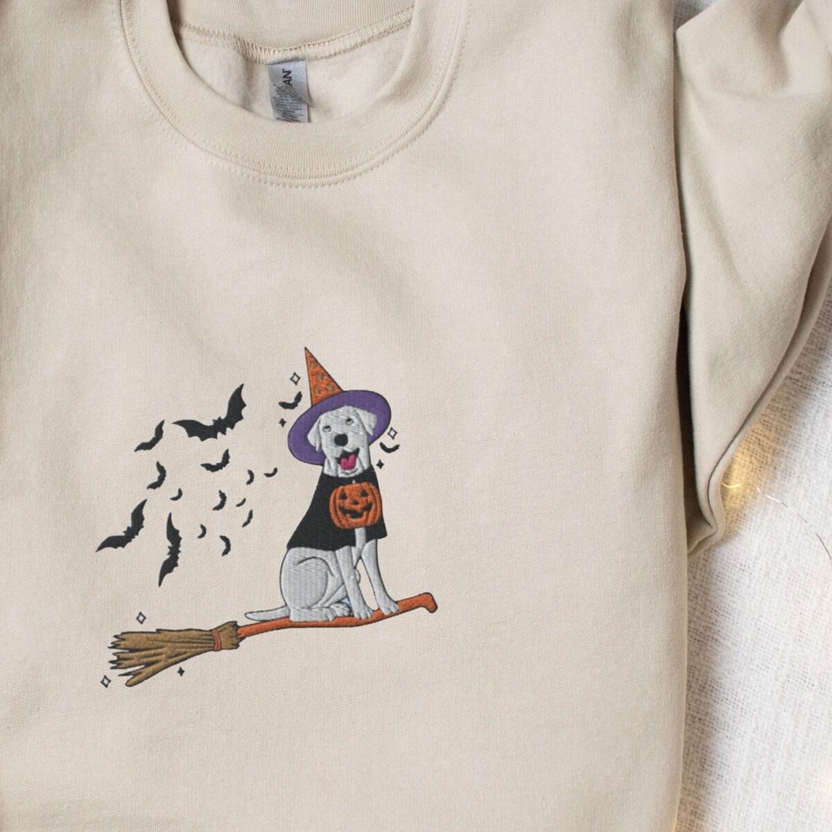 Labrador Witch Sweatshirt – Sand Sand crewneck sweatshirt laid flat with a sleeve folded over, embroidered with a pale cream lab in a purple and orange witch hat and a jack-o'-lantern cape, riding a broomstick with a trail of bats