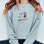 Woman with a gold necklace in a Sport Grey crewneck sweatshirt embroidered with a pale cream lab in a purple and orange witch hat and a jack-o'-lantern cape, riding a broomstick with a trail of bats