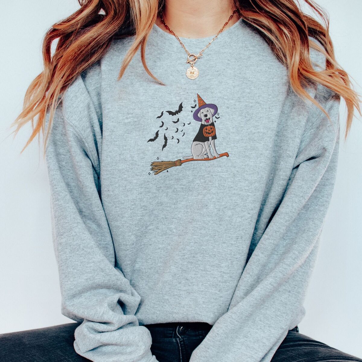 Labrador Witch Sweatshirt - Sport Grey Woman with a gold necklace in a Sport Grey crewneck sweatshirt embroidered with a pale cream lab in a purple and orange witch hat and a jack-o'-lantern cape, riding a broomstick with a trail of bats