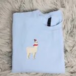 Llama Christmas sweatshirt in light blue with a small embroidered cream llama wearing a red pom-pom beanie and a red scarf