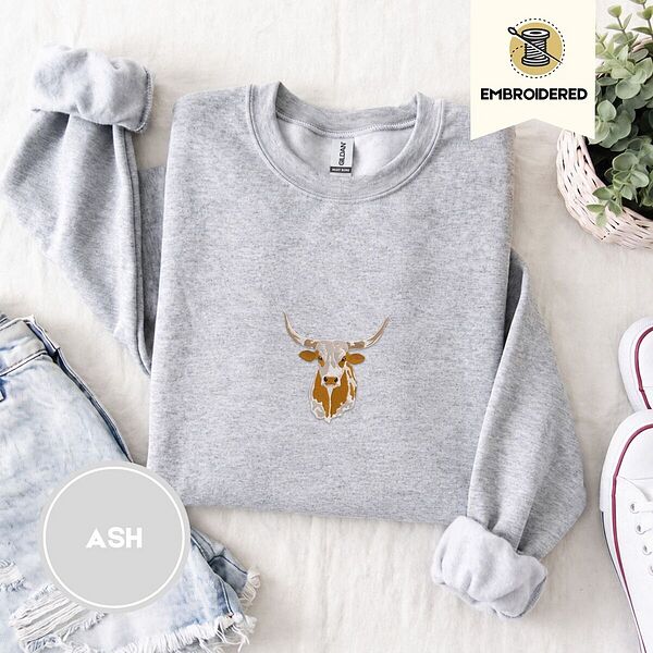 Ash crewneck sweatshirt folded with denim shorts, white sneakers and a potted plant, embroidered with a longhorn steer head with cream and copper patches and long curved horns