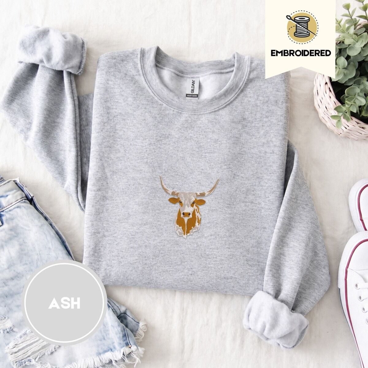 Longhorn Cow Sweatshirt – Ash Ash crewneck sweatshirt folded with denim shorts, white sneakers and a potted plant, embroidered with a longhorn steer head with cream and copper patches and long curved horns