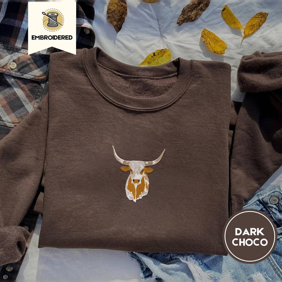 Longhorn Cow Sweatshirt – Dark Chocolate Dark Chocolate crewneck sweatshirt on a plaid flannel shirt with yellow autumn leaves and denim shorts, embroidered with a longhorn steer head with cream and copper patches and long curved horns