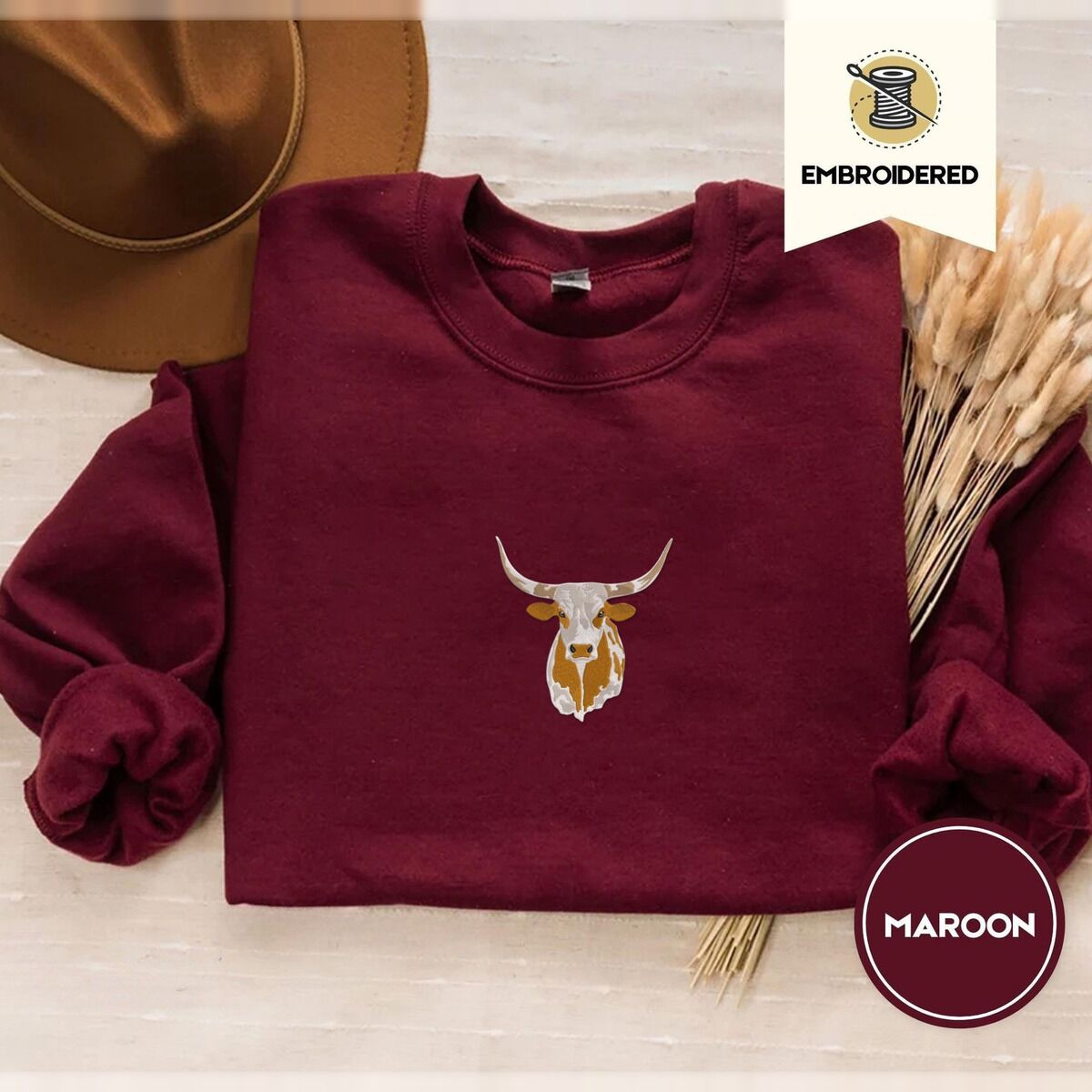 Longhorn Cow Sweatshirt – Maroon Maroon crewneck sweatshirt with a brown felt hat and dried bunny tail grass, embroidered with a longhorn steer head with cream and copper patches and long curved horns