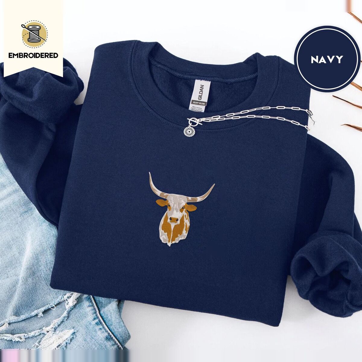 Longhorn Cow Sweatshirt – Navy Navy crewneck sweatshirt folded with ripped jeans and a silver chain necklace, embroidered with a longhorn steer head with cream and copper patches and long curved horns
