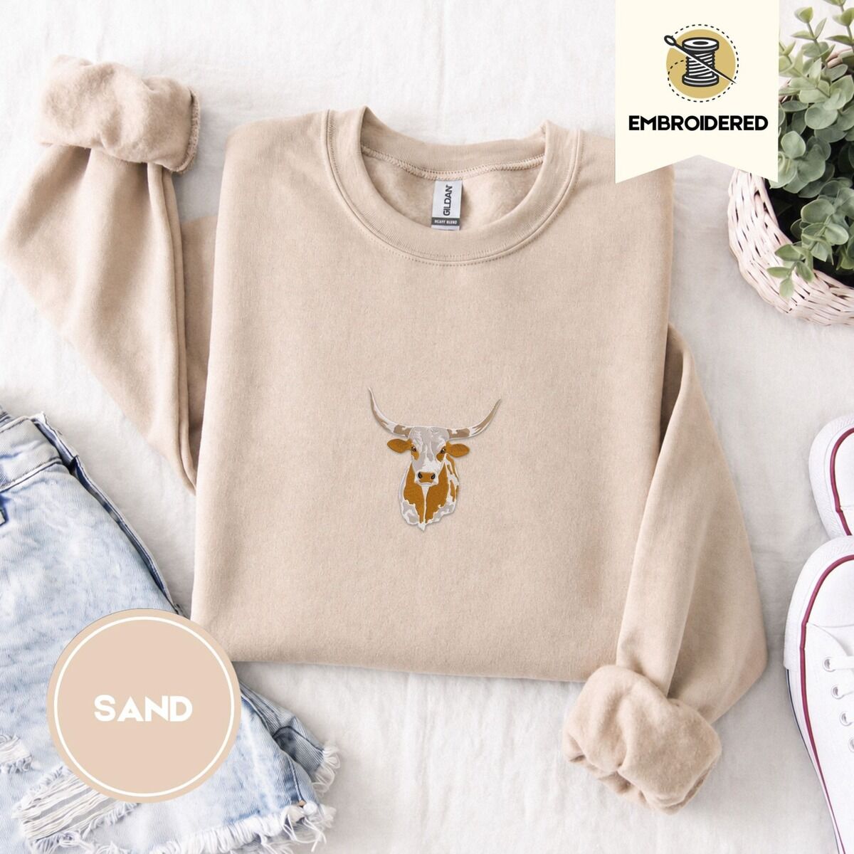 Longhorn Cow Sweatshirt – Sand Sand crewneck sweatshirt folded with denim shorts, white sneakers and a potted plant, embroidered with a longhorn steer head with cream and copper patches and long curved horns