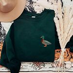 Forest Green crewneck sweatshirt laid flat on a printed blanket with dried pampas grass, embroidered with a standing drake mallard with a green head, orange bill, chestnut chest and grey wing on the left chest