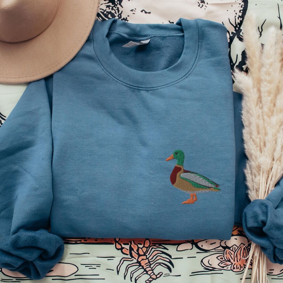 Mallard Crewneck – Indigo Blue Indigo Blue crewneck sweatshirt laid flat beside a felt hat and pampas grass, embroidered with a standing drake mallard with a green head, orange bill, chestnut chest and grey wing on the left chest