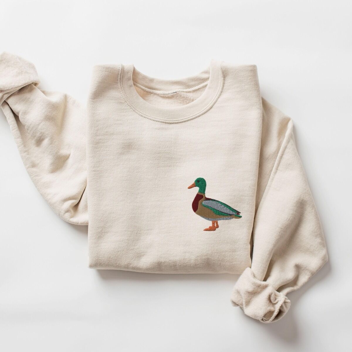 Mallard Crewneck – Sand Sand crewneck sweatshirt laid flat with a rolled cuff, embroidered with a standing drake mallard with a green head, orange bill, chestnut chest and grey wing on the left chest