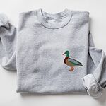Sport Grey crewneck sweatshirt laid flat with a rolled cuff, embroidered with a standing drake mallard with a green head, orange bill, chestnut chest and grey wing on the left chest