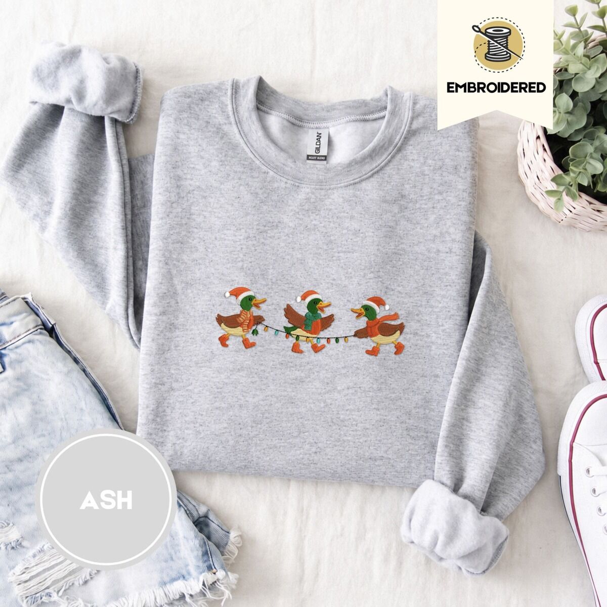 Mallard Duck Christmas Sweatshirt – Ash Ash crewneck sweatshirt folded with denim shorts, white sneakers and a potted plant, embroidered with three mallard ducks in Santa hats, knit scarves and orange boots carrying a string of Christmas lights