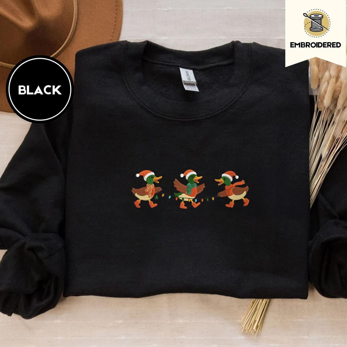 Mallard Duck Christmas Sweatshirt – Black Black crewneck sweatshirt with a brown felt hat and dried bunny tail grass, embroidered with three mallard ducks in Santa hats, knit scarves and orange boots carrying a string of Christmas lights