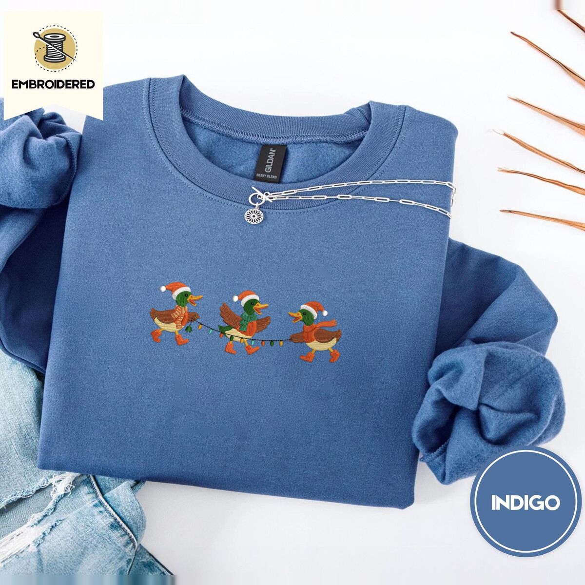 Mallard Duck Christmas Sweatshirt – Indigo Blue Indigo Blue crewneck sweatshirt folded with light jeans and dried twigs, embroidered with three mallard ducks in Santa hats, knit scarves and orange boots carrying a string of Christmas lights