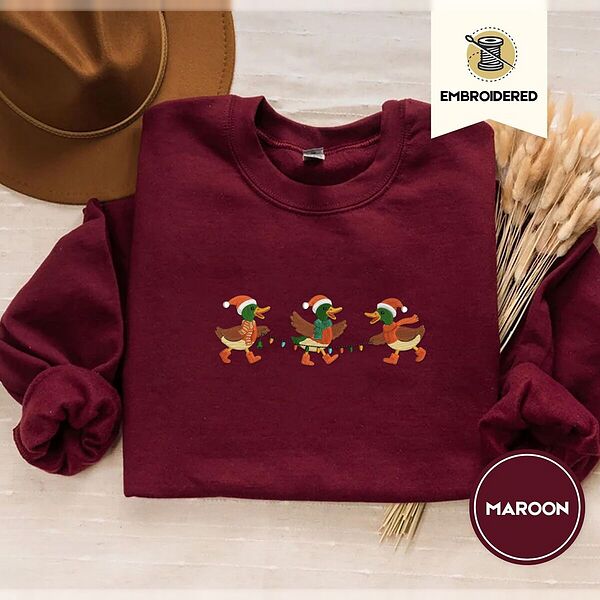 Maroon crewneck sweatshirt with a brown felt hat and dried bunny tail grass, embroidered with three mallard ducks in Santa hats, knit scarves and orange boots carrying a string of Christmas lights